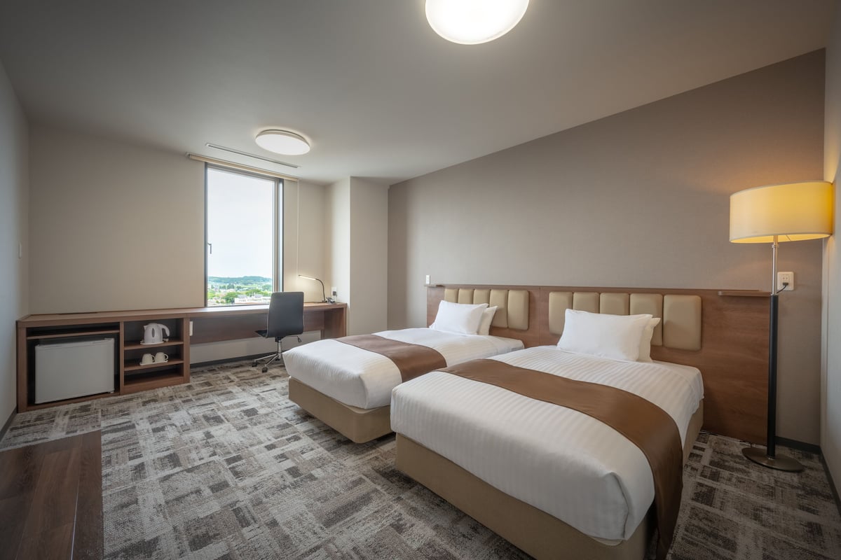 Ett hotellrum med två sängar, ett skrivbord, en stol, en lampa och utsikt över omgivningarna.
