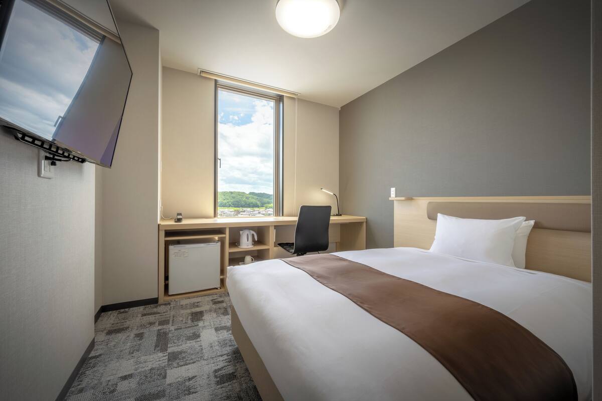 Ett hotellrum med en säng, ett skrivbord med en stol, en tv och ett fönster med utsikt över grönområden.