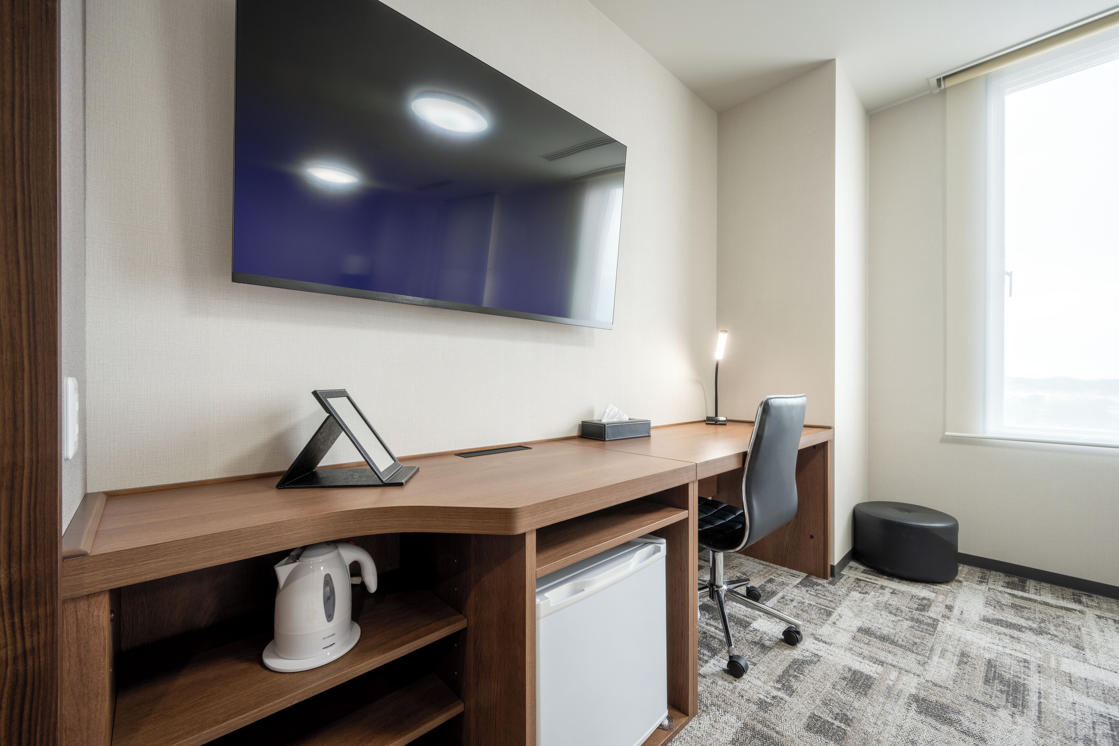 A modern office space with a large flat-screen TV mounted on the wall, a wooden desk with a kettle, a trash bin, and a chair.