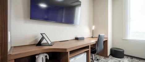 A modern office space with a large flat-screen TV mounted on the wall, a wooden desk with a kettle, a trash bin, and a chair.
