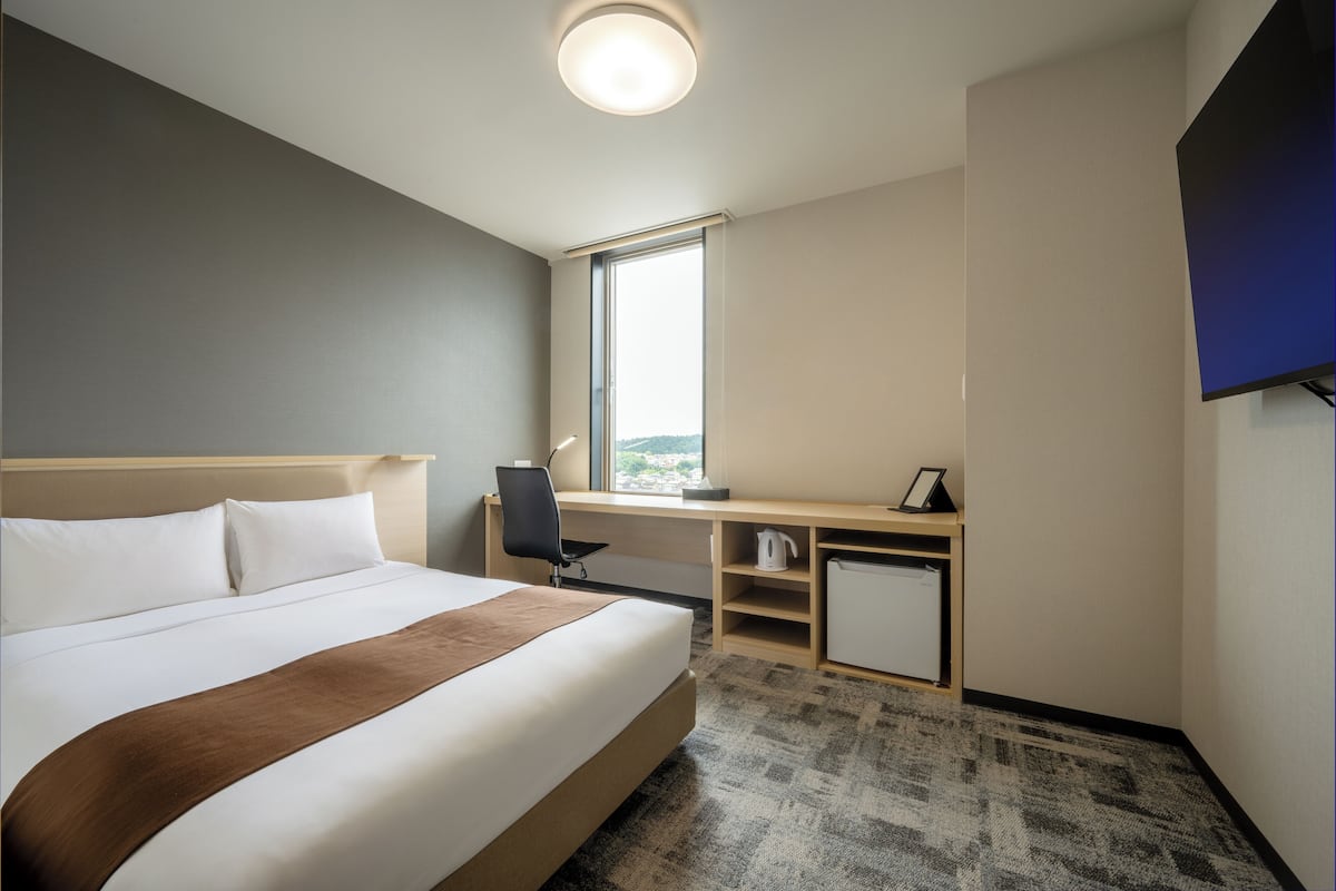 Ett hotellrum med en stor säng, ett skrivbord, en stol, ett litet skåp och ett stort fönster.