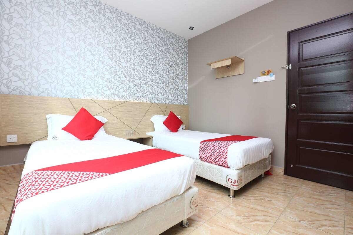 Een hotelkamer met twee bedden, elk met rode en witte beddengoed, een houten hoofdbord, een patroon op de muur, een klein plankje en een donker houten deur.