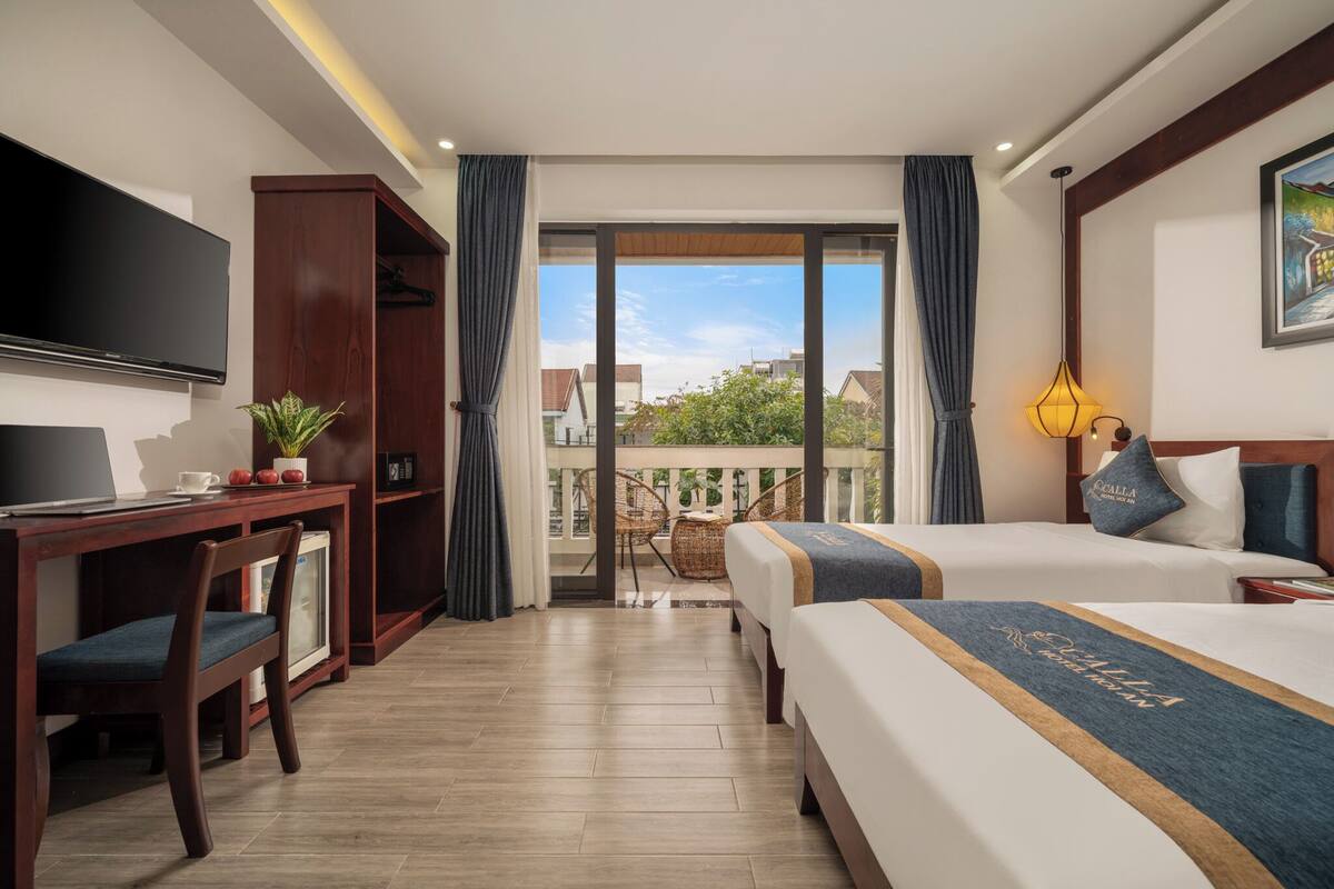 A hotel room with two beds, a desk with a TV, a small fridge, a balcony with a view of buildings and trees.