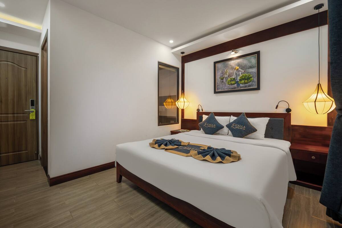 A neatly made bed with a white comforter and blue pillows, a painting on the wall, and two wall-mounted lamps.