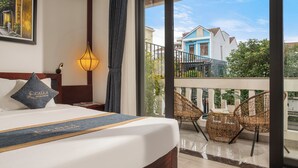 A hotel room with a large bed, a balcony with wicker chairs, and a view of neighboring houses. - Calla Hotel Hoi an (Da Nang)