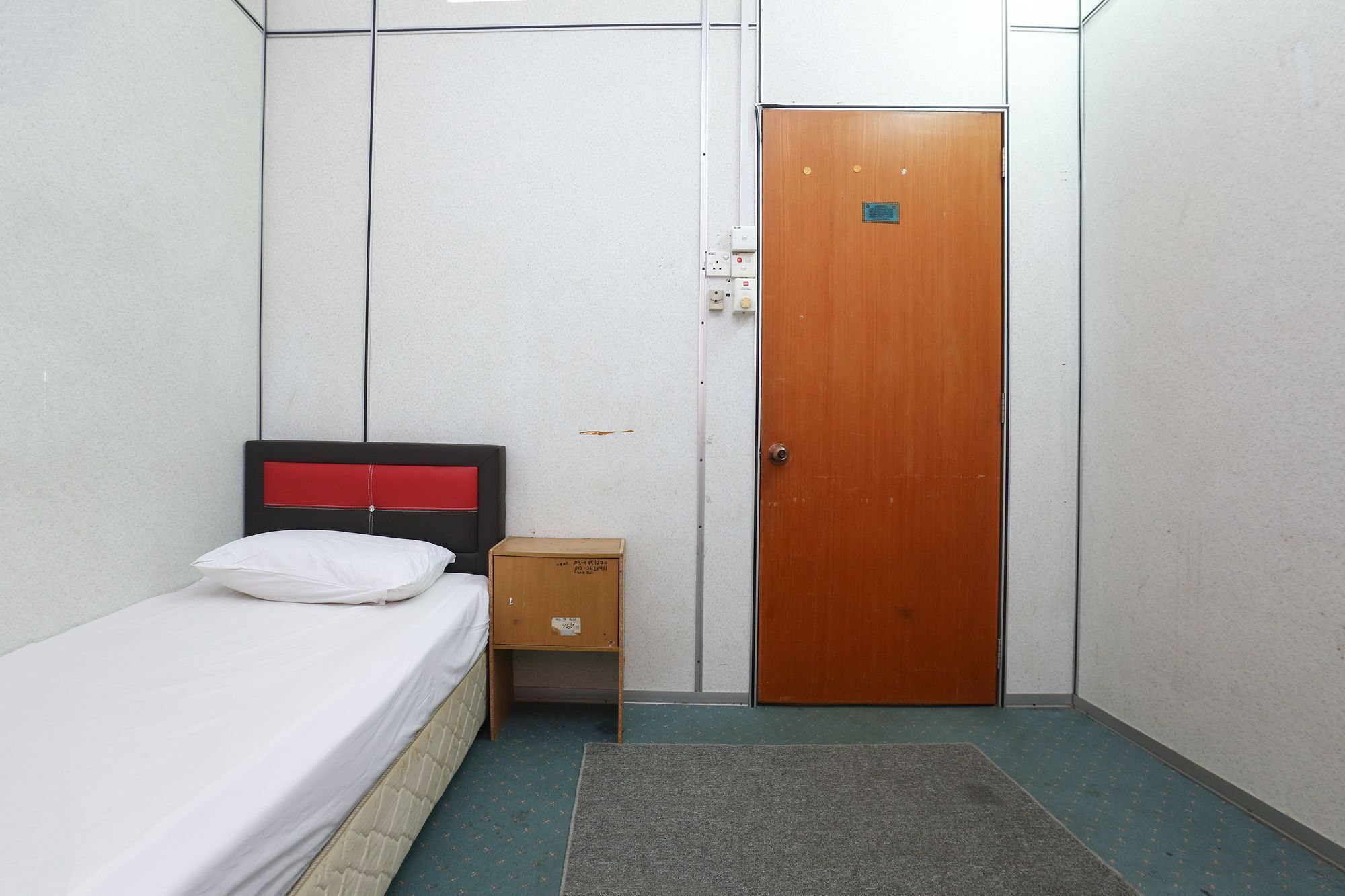 A small, single-bed room with a wooden door, a small wooden cabinet, and a grey carpet.