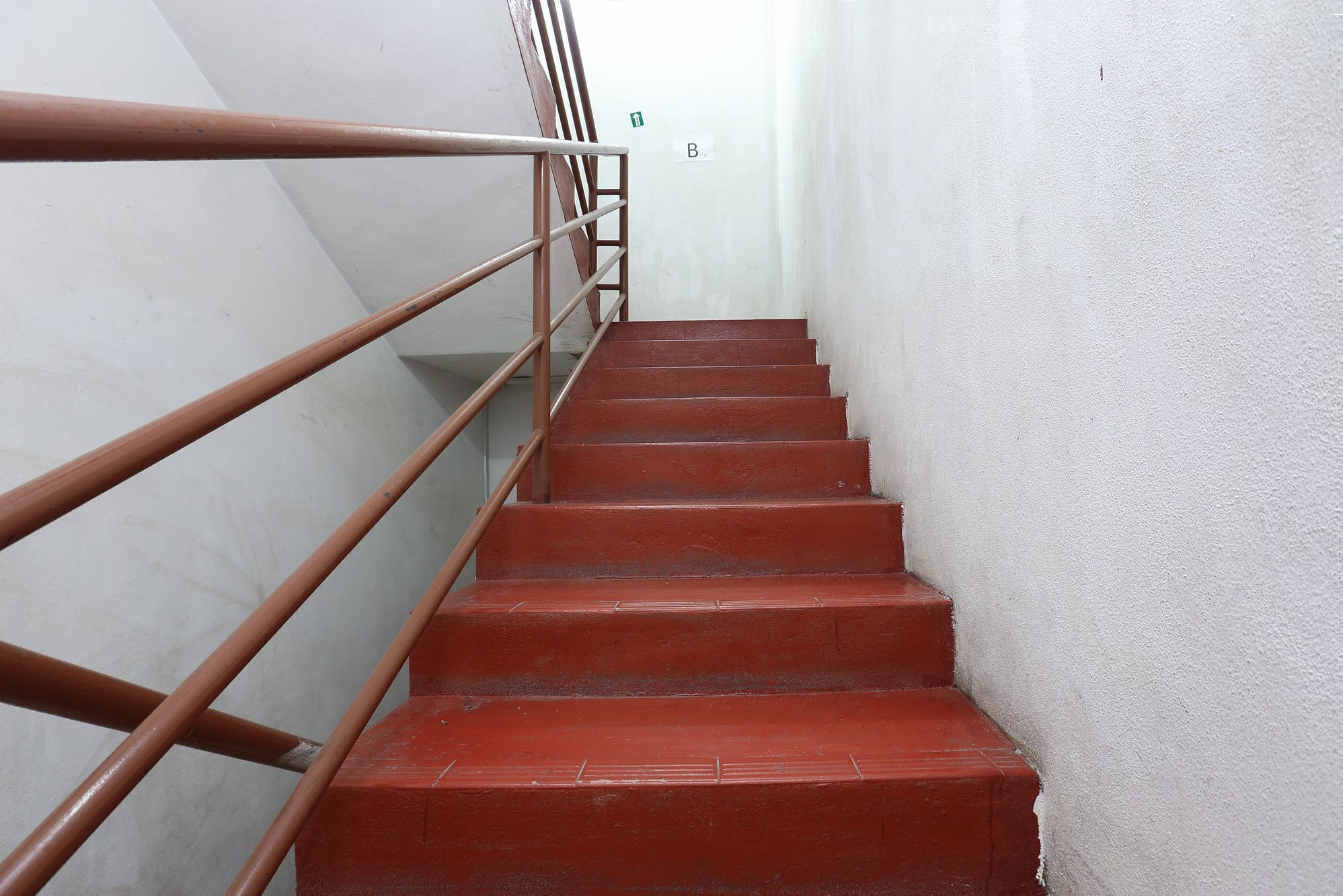 A staircase with red steps and a metal handrail, leading to a white wall with a green exit sign.