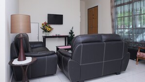 Una sala de estar con dos sofás de cuero negro, una mesita redonda con una lámpara, un televisor montado en la pared y un ventanal con cortinas.