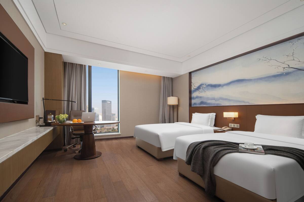 A hotel room with two beds, a desk with a laptop, a TV, and a large window with a city view.