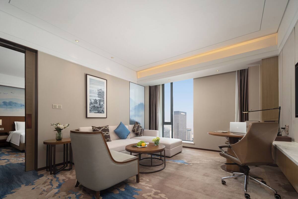 A modern hotel room with a sofa, armchair, round coffee table, and a desk with a laptop. Large windows offer a city view.