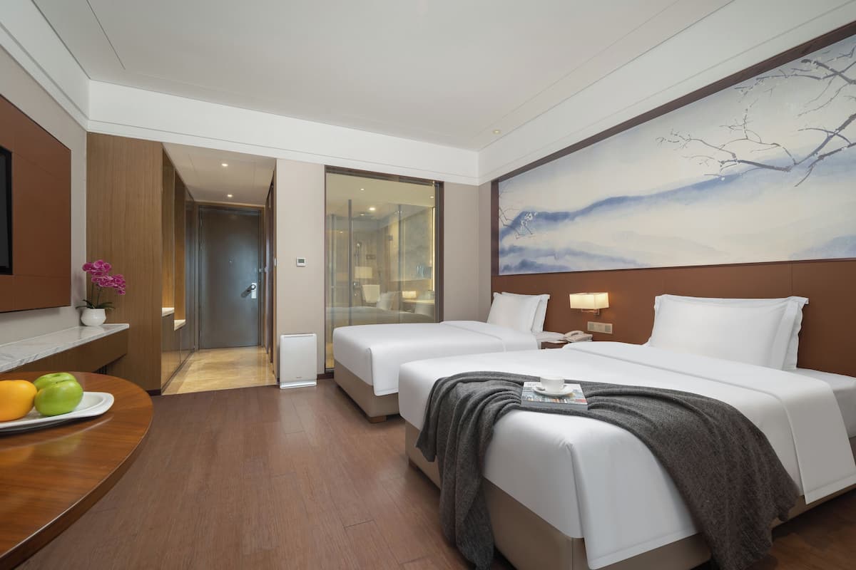 A hotel room with two beds, a wooden desk with a fruit plate, a painting on the wall, and a view of the hallway.
