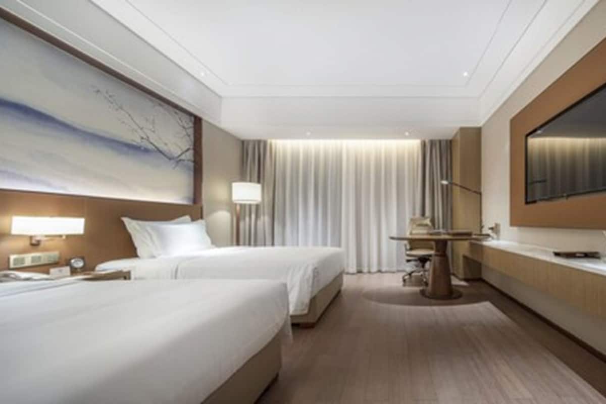 A hotel room with two beds, a desk, a TV, and a large window with curtains.