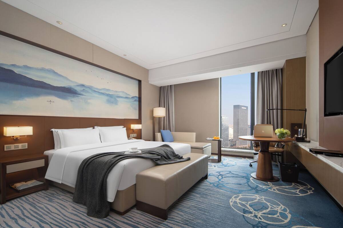 A hotel room with a large bed, a desk with a laptop, a chair, a small table with a plant, and a large window with a city view.