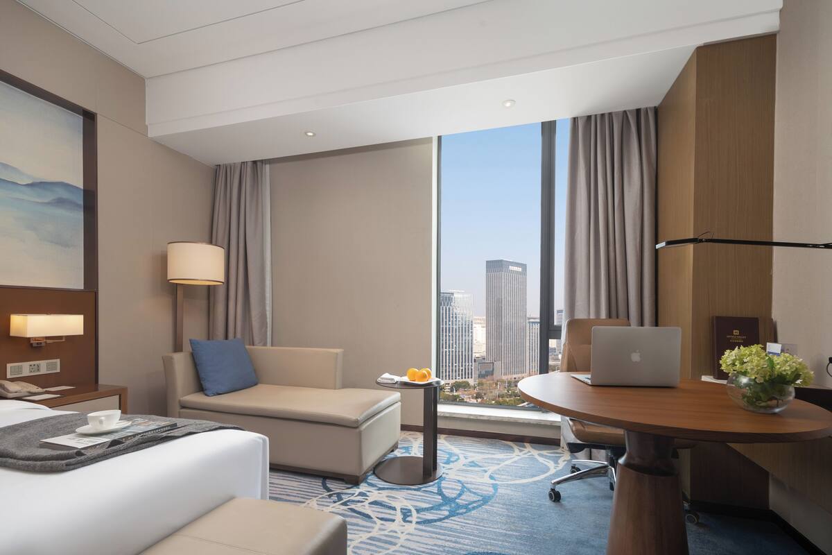 A modern hotel room with a bed, a sofa, a desk with a laptop, a small table with a vase, and a large window with a city view.