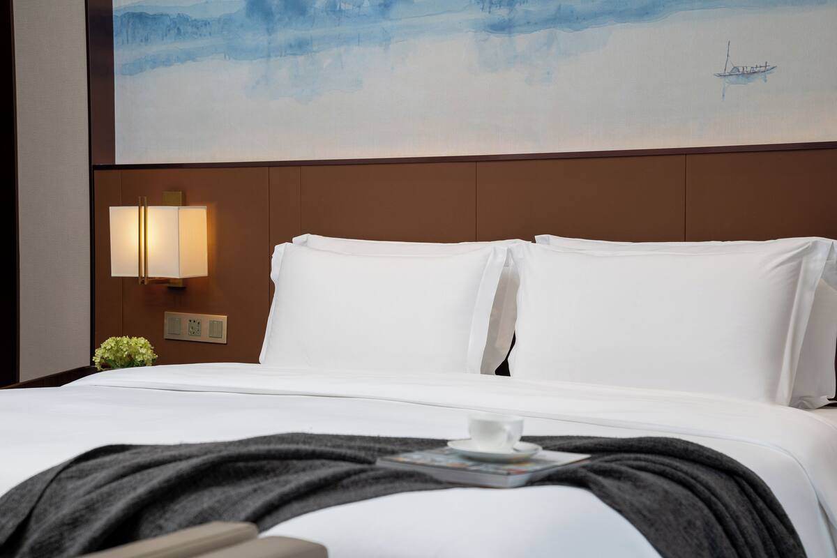 A hotel room with a neatly made bed, a painting of a boat on the wall, a bedside lamp, and a small green plant.