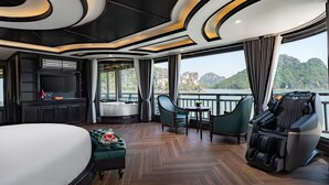 A luxurious suite with a large bed, a massage chair, a seating area with a table, a TV, and a scenic view of mountains and water. - Rita Cruise (Tuan Chau)