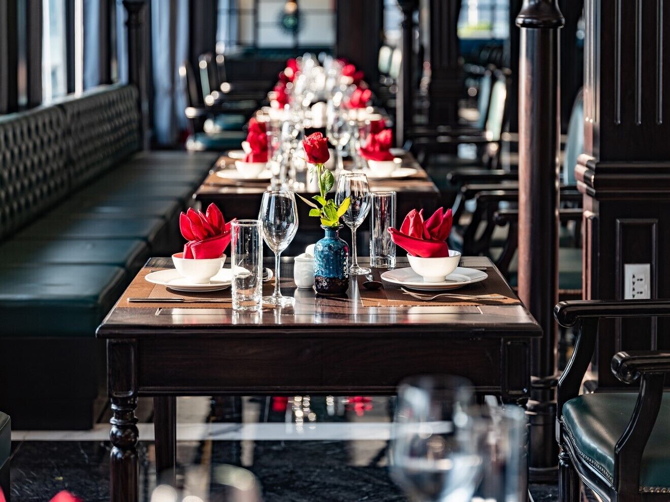 A long dining table set with red napkins, clear glasses, and a blue vase with a red rose.