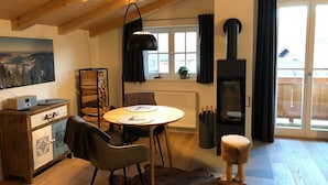 A cozy room with a round wooden table, two chairs, a small stool, a fireplace, and a large window with curtains. - Bodenschneid Suites Neureuthblick - Bodenschneid Suite Neureuthblick (Kreuth)