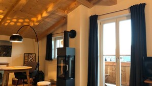 A cozy room with a wooden ceiling, a dining table, chairs, a black heater, and a large sliding glass door with curtains. - Bodenschneid Suites Neureuthblick - Bodenschneid Suite Neureuthblick (Kreuth)