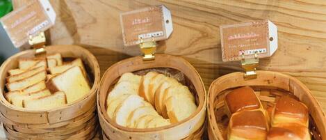 Três cestas de madeira com produtos de panificação, cada uma com etiqueta de preço.