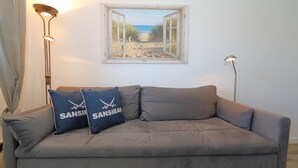 A grey sofa with two blue pillows, a framed beach view, and two floor lamps. - Tanja Hüs - in the south grove (Sylt)