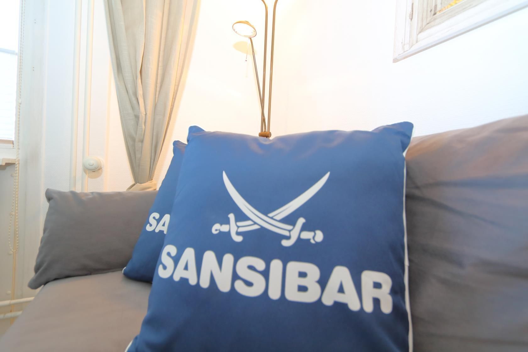 A blue pillow with "SANSIBAR" and a sword design on a sofa.