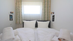 A neatly made bed with white linens and pillows, a window with curtains, and two wall decorations. - Tanja Hüs - in the south grove (Sylt)