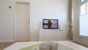 A room with a television on a cabinet, a door, and a window with curtains. - Tanja Hüs - in the south grove (Sylt)