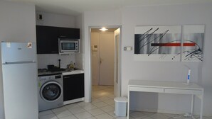 A compact kitchen with a refrigerator, microwave, washing machine, and sink. There's a small table with a lamp and a trash bin. - One-Bedroom Apartment, good standard, quiet with parking (Lyon)