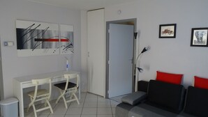 A modern living room with a white desk, two chairs, a black sofa with red cushions, and artwork on the walls. - One-Bedroom Apartment, good standard, quiet with parking (Lyon)