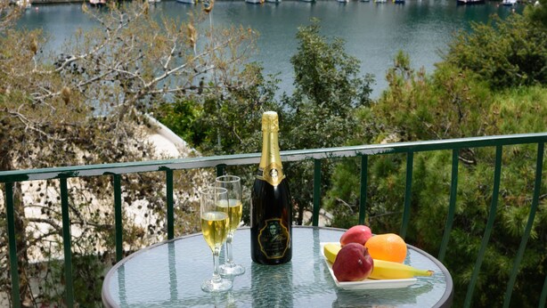 Una mesa con una botella de champán, dos copas y un plato de fruta en un balcón con vistas al agua y al puerto deportivo.