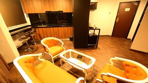 A modern kitchen with a black countertop, wooden cabinets, and a black stove. There are three orange chairs with white frames and a small white table. - Golden Nasmir Hotel (Perai)