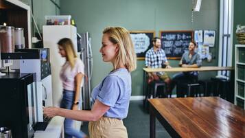 Een coffeeshop met een barista die drankjes bereidt achter de bar, een klant op de achtergrond en een zitgedeelte met tafels.
