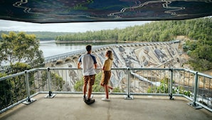 A couple standing on a concrete platform overlooking a dam with a river and forested hills in the background. - Black Diamond Lodge (Collie)