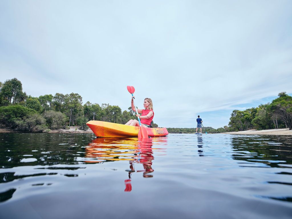 Une personne pratiquant le kayak sur une rivière paisible, avec des arbres et un ciel dégagé en arrière-plan.
