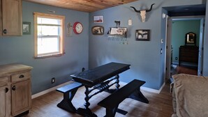 Une pièce avec un plafond en bois, une table à manger avec des bancs, une fenêtre avec des stores et des éléments de décoration murale, notamment une horloge, un crâne de vache et des tableaux encadrés.