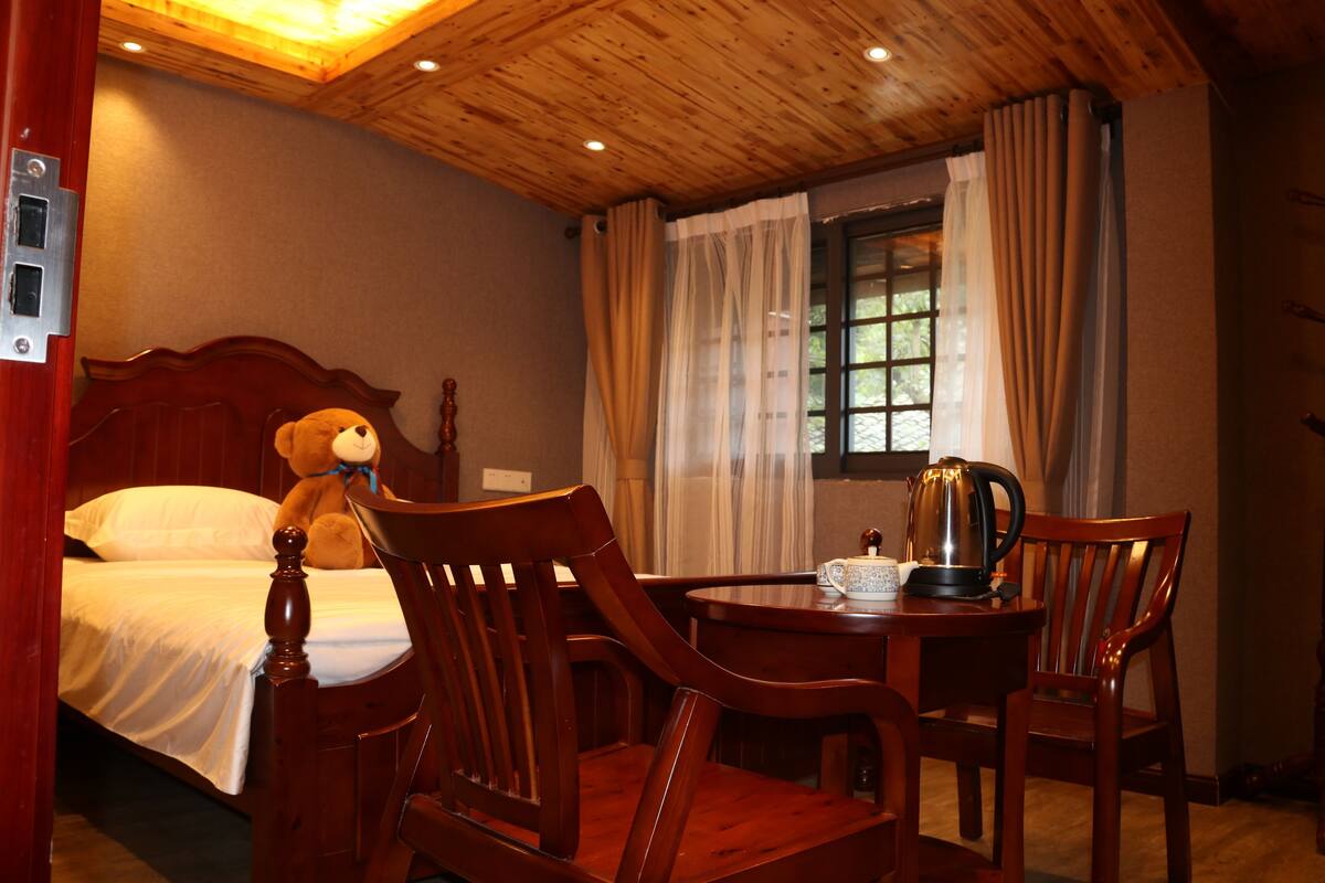 A hotel room with a wooden ceiling, a bed with a teddy bear, a dining table with chairs, and a window with curtains.