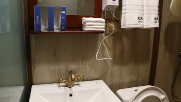A bathroom with a sink, a mirror, a towel dispenser, and a shelf with toiletries.