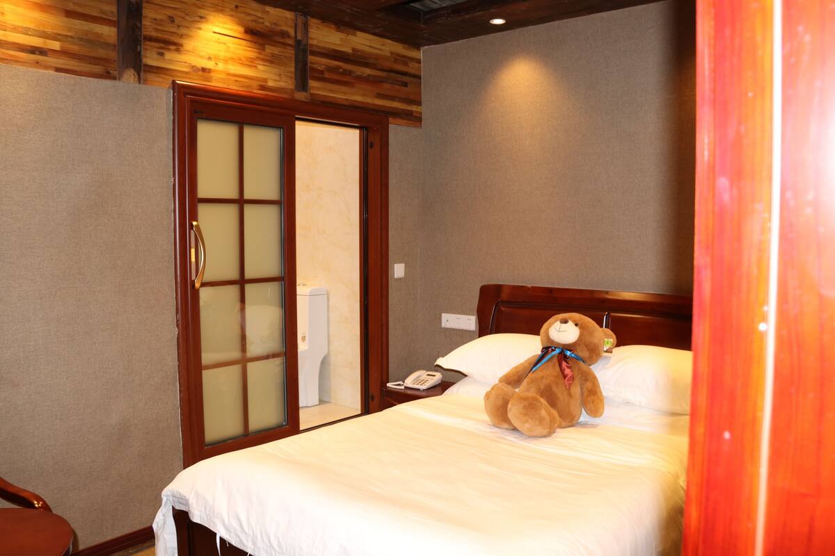 A hotel room with a bed, a teddy bear, a wooden door, and a telephone on the nightstand.