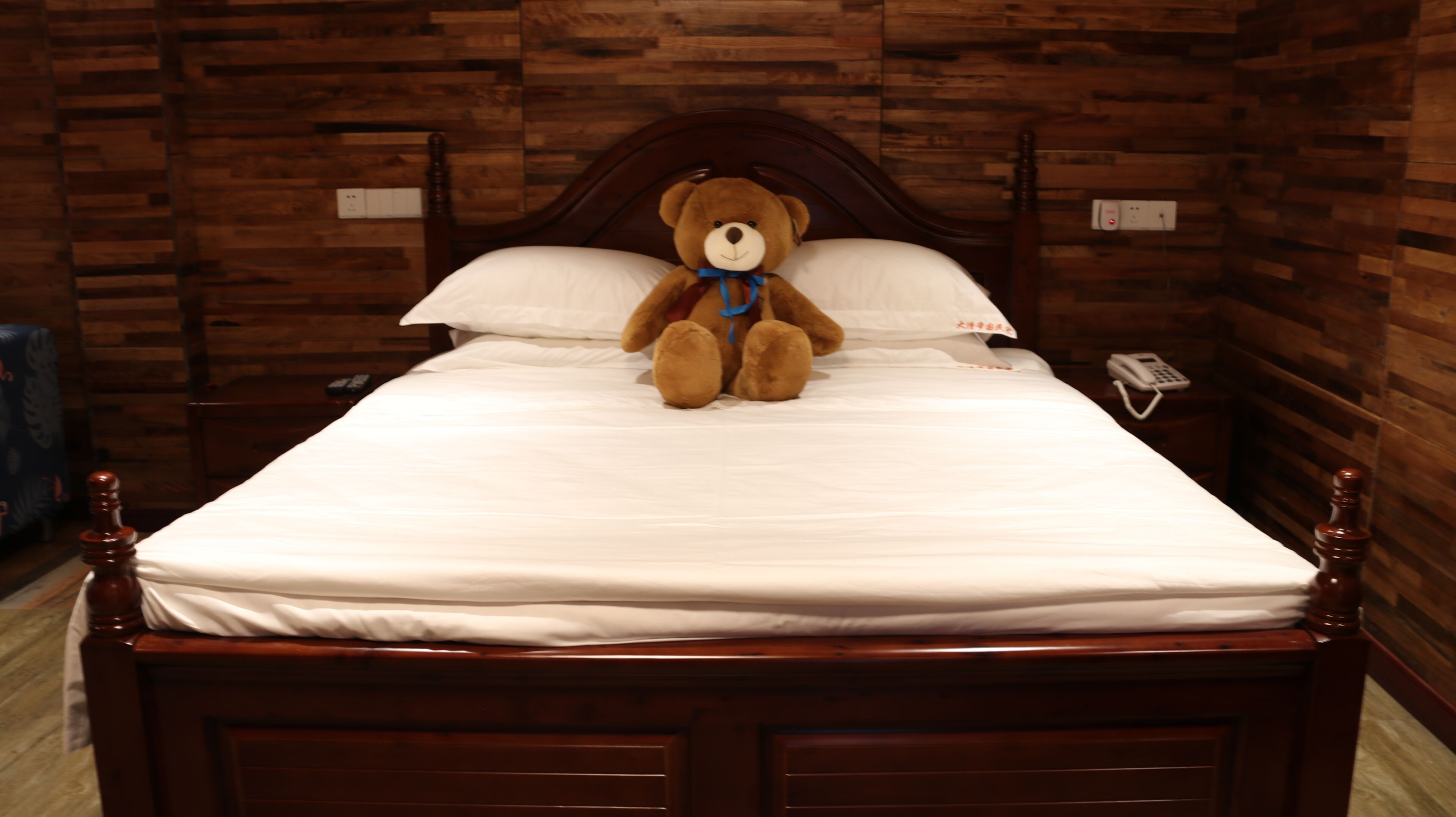 Una cama con un osito de peluche, cabecera de madera, ropa de cama blanca y un teléfono en la mesita de noche.