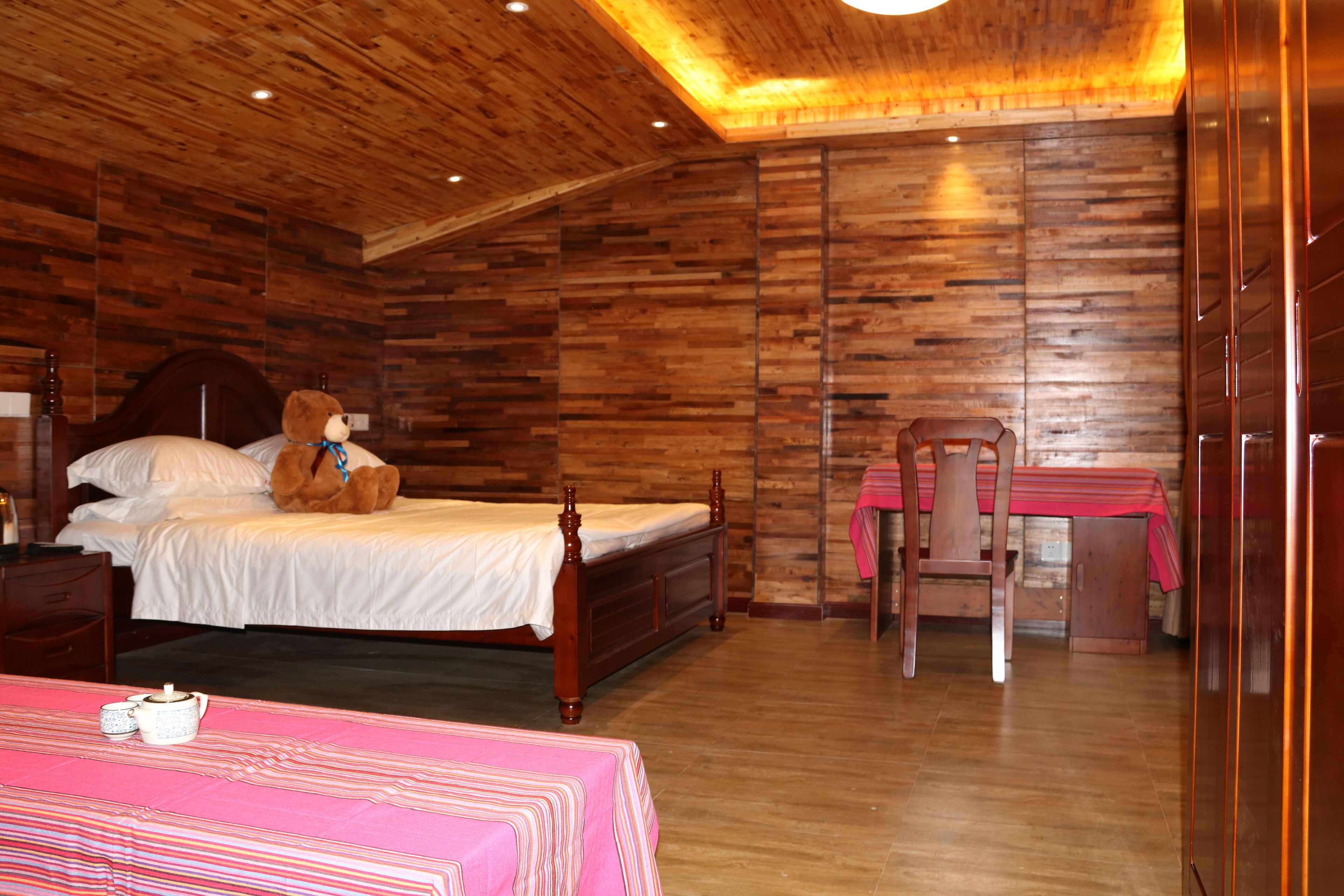 Un dormitorio con techo de madera, una cama con un oso de peluche, una mesita con mantel rojo y una silla.