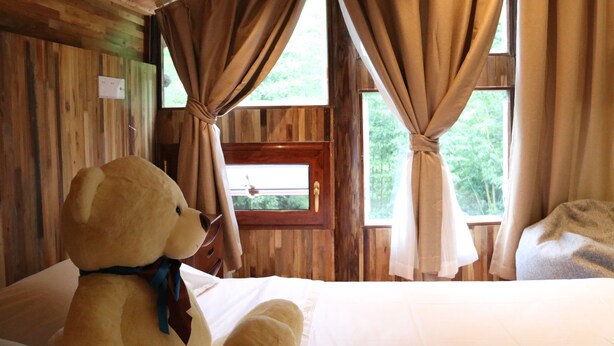 A wooden cabin room with a bed, a window with curtains, and a teddy bear on the bed. - Qing Dynasty House (Leshan)