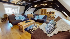 Een zolderkamer met zichtbare houten balken, twee banken met gestreepte kussens, een houten salontafel en een eethoek met houten stoelen en een tafel.