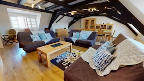 An attic room with exposed wooden beams, two sofas with patterned cushions, a wooden coffee table, and a dining area with wooden chairs and a table.
