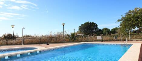 Une piscine rectangulaire à l’eau d’un bleu limpide, entourée d’un petit muret en briques et d’une clôture en métal. Deux lampadaires sont disposés le long de la clôture, et on aperçoit un arbre au fond.