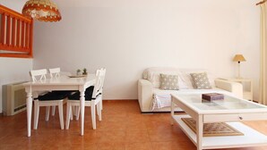 Una sala de estar con una mesa de comedor, sillas blancas, un sofá con cojines estampados, una mesa de centro de vidrio y una lámpara colgante.