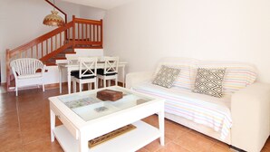 Una sala de estar con un sofá blanco, una mesa de centro de vidrio, una silla de mimbre blanca, una escalera de madera y una mesa de comedor con sillas.