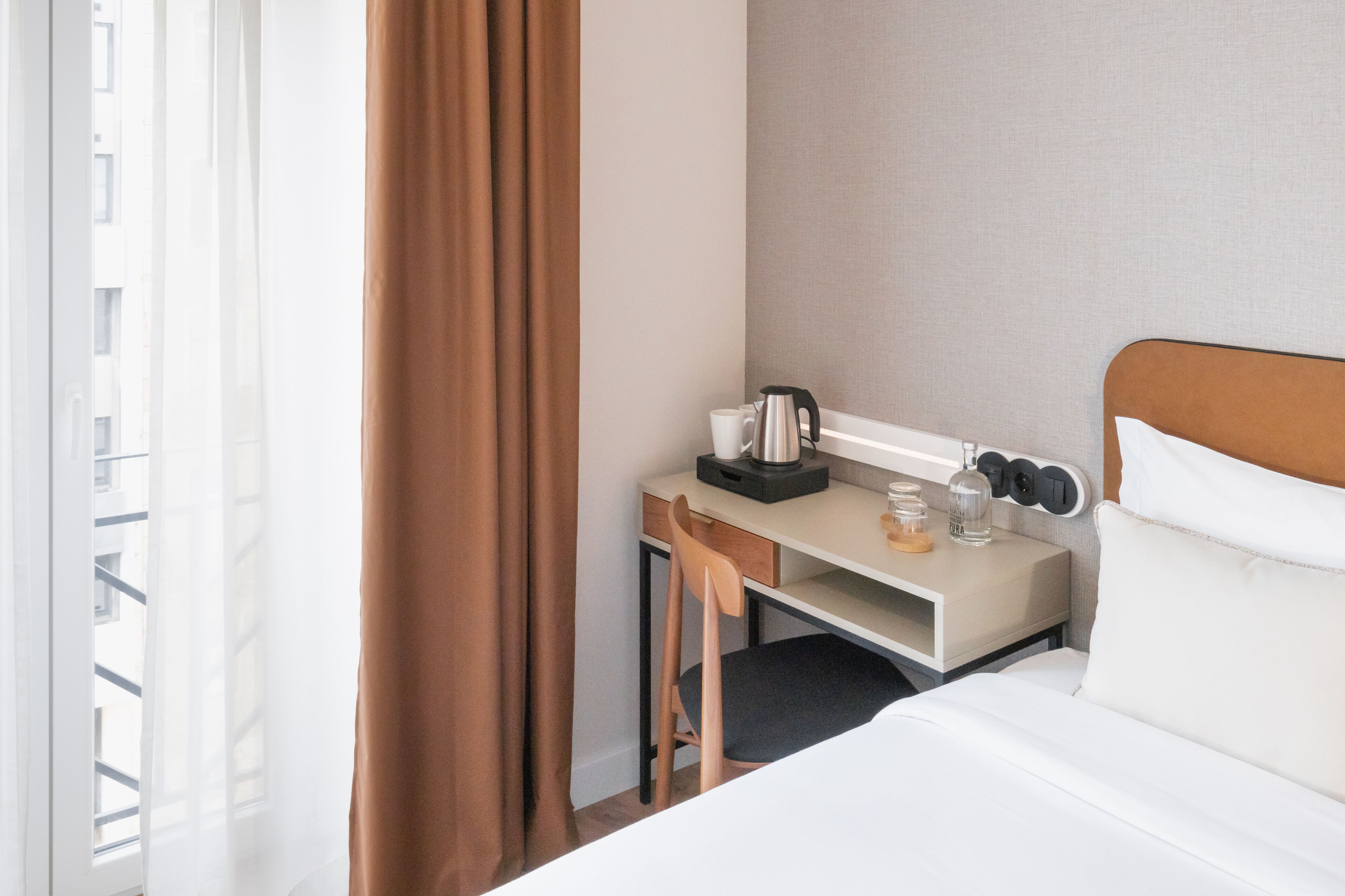 A hotel room with a bed, a desk with a coffee maker, a chair, and a nightstand with a lamp.