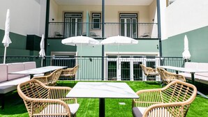 An outdoor patio with wicker furniture and white umbrellas, a green lawn, and a building with balconies in the background. - Lx SoHo Boutique Hotel by RIDAN Hotels (Lisbon)