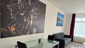 A modern living room with a glass dining table, black leather chairs, a grey sofa, and a large cityscape painting on the wall.